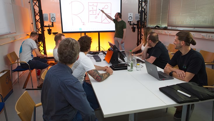 Several young people sit at a table, looking at a smartboard where a model is being explained.