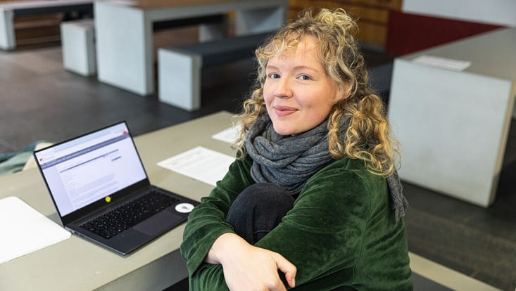 A young woman smiles and looks at the camera, with her laptop visible on the table in front of her.