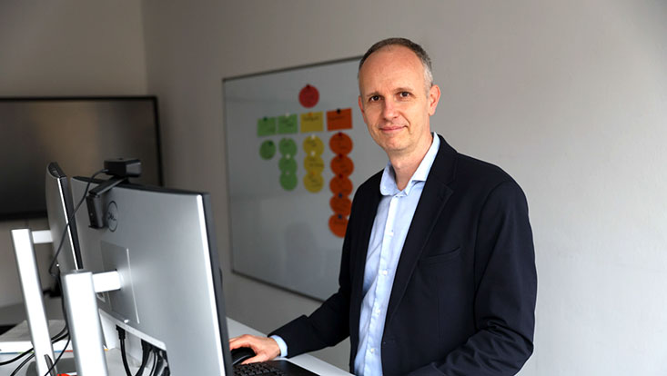 A man stands at his workstation, with a whiteboard covered in Post-its in the background.